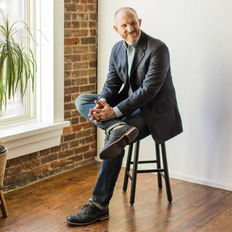 photo of Simon Trask sitting on a stool next to a window and brick wall